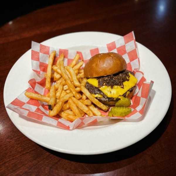 A cheeseburger with caramelized onions, fries, and pickles on a white plate with red checkered paper.