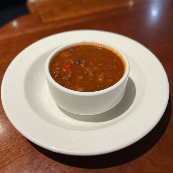 A white bowl of chili with beans and red peppers sits on a white plate on a wooden table.