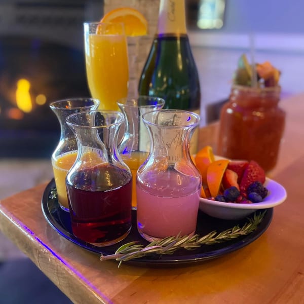 Mimosa bar with various juices, fruit, and a bottle of champagne on a wooden table.