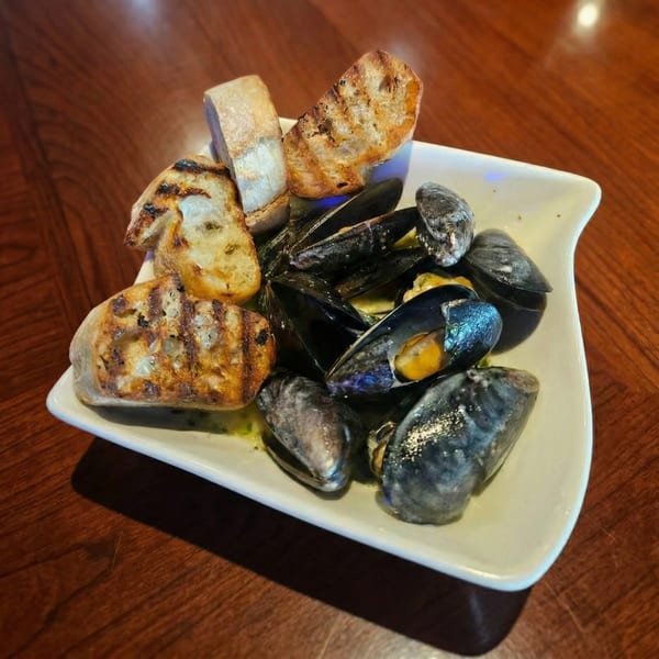 A white bowl of mussels in broth with grilled bread on a wooden table.